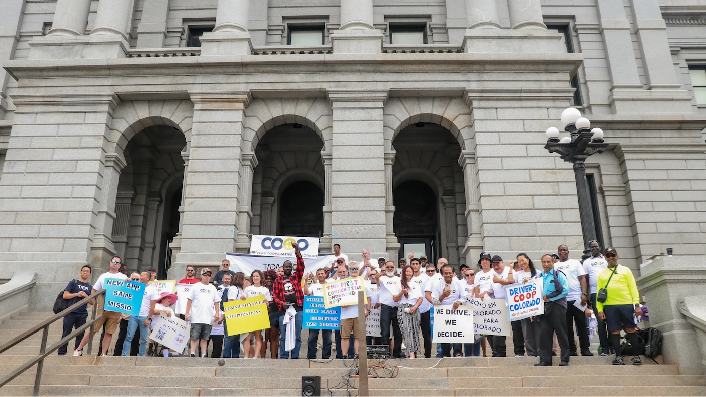 Group of people gathered on the steps of a government building holding signs in support of Drivers Cooperative Colorado, advocating for driver-owned rideshare alternatives.