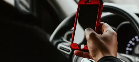 Close-up of a person holding a smartphone with a red case while sitting behind the wheel of a car, illustrating distracted driving.
