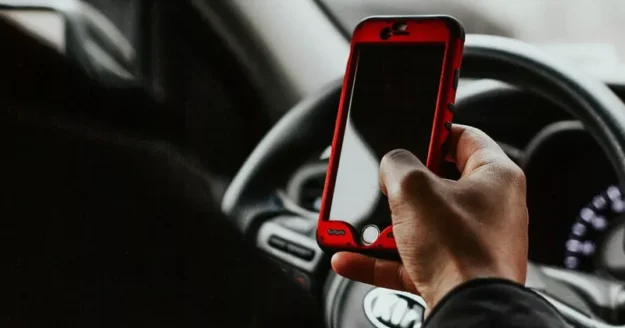 Close-up of a person holding a smartphone with a red case while sitting behind the wheel of a car, illustrating distracted driving.